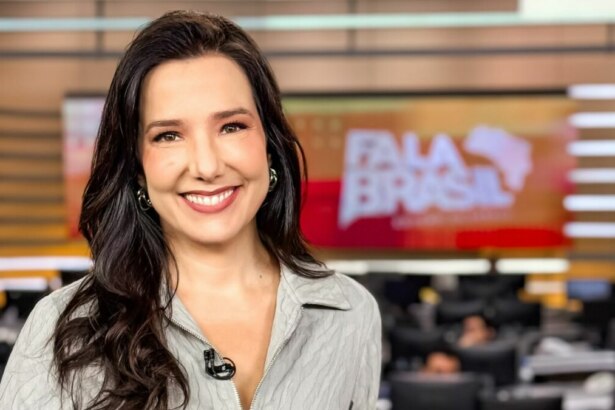 Adriana Perroni, jornalista do programa "Fala Brasil", sorri para a câmera em um estúdio de telejornal. Ela tem cabelos escuros e ondulados e usa uma camisa cinza clara. Ao fundo, pode-se ver o logo do "Fala Brasil" em um telão e o ambiente de uma redação.