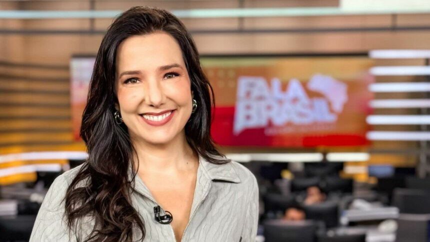 Adriana Perroni, jornalista do programa "Fala Brasil", sorri para a câmera em um estúdio de telejornal. Ela tem cabelos escuros e ondulados e usa uma camisa cinza clara. Ao fundo, pode-se ver o logo do "Fala Brasil" em um telão e o ambiente de uma redação.