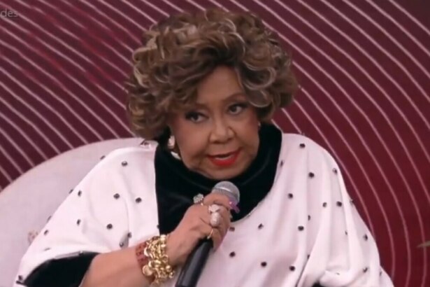 Alcione, com cabelo volumoso e maquiagem destacada, usando um casaco branco com bolinhas pretas e detalhes em veludo preto, segura um microfone e olha diretamente para a câmera. Ela está sentada em um sofá branco, com um fundo desfocado em tons de vermelho e rosa, durante uma participação no programa "É de Casa".