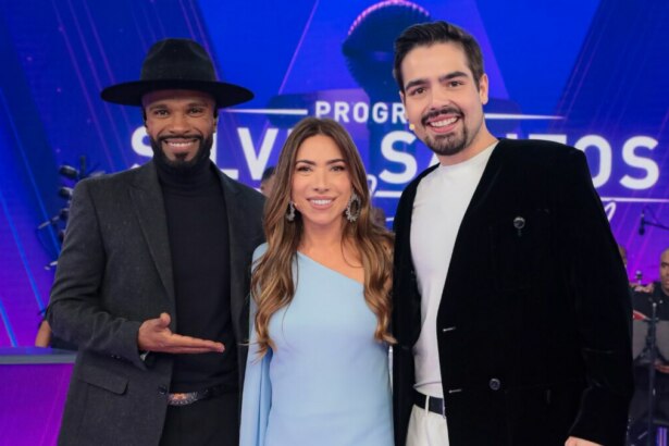 Alexandre Pires (esquerda, de chapéu e roupa escura), Patrícia Abravanel (centro, de vestido azul claro) e João Silva (direita, de blazer preto e camiseta branca) posam sorridentes no palco do "Programa Silvio Santos". Ao fundo, o logo do programa e músicos são visíveis.