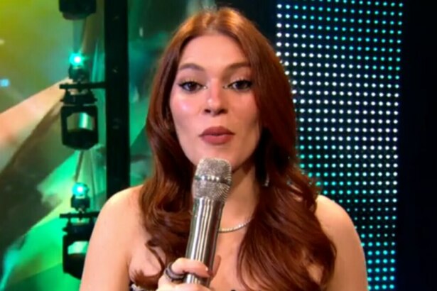 A imagem é um close da apresentadora Ana Clara, sorrindo e falando em um microfone no palco do Estrela da Casa. Ela está usando um vestido brilhante e um colar prateado. O cabelo dela, castanho avermelhado, está solto, no palco do Estrela da Casa