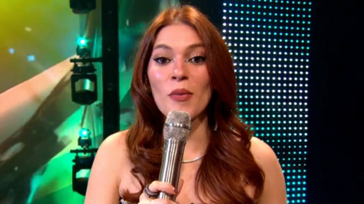 A imagem é um close da apresentadora Ana Clara, sorrindo e falando em um microfone no palco do Estrela da Casa. Ela está usando um vestido brilhante e um colar prateado. O cabelo dela, castanho avermelhado, está solto, no palco do Estrela da Casa