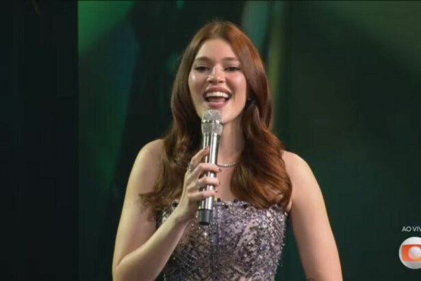 A imagem é um close da apresentadora Ana Clara, sorrindo e falando em um microfone no palco do Estrela da Casa. Ela está usando um vestido brilhante e um colar prateado. O cabelo dela, castanho avermelhado, está solto. Ao fundo, um cenário verde e escuro, com o logo da TV Globo no canto inferior direito.