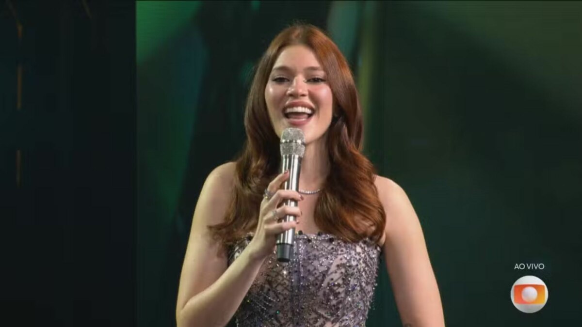 A imagem é um close da apresentadora Ana Clara, sorrindo e falando em um microfone no palco do Estrela da Casa. Ela está usando um vestido brilhante e um colar prateado. O cabelo dela, castanho avermelhado, está solto. Ao fundo, um cenário verde e escuro, com o logo da TV Globo no canto inferior direito.