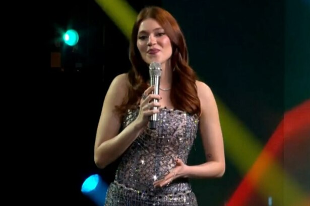 A imagem é da apresentadora Ana Clara, sorrindo e falando em um microfone no palco do Estrela da Casa. Ela está usando um vestido brilhante e um colar prateado. O cabelo dela, castanho avermelhado, está solto, no palco do Estrela da Casa