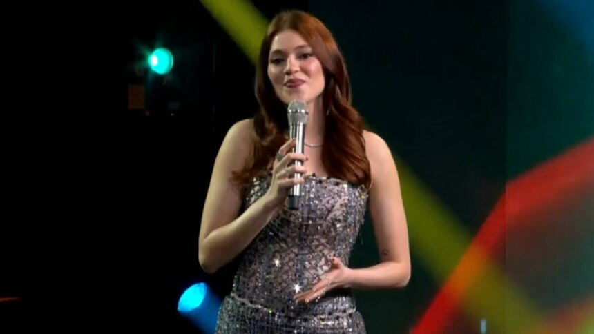 A imagem é da apresentadora Ana Clara, sorrindo e falando em um microfone no palco do Estrela da Casa. Ela está usando um vestido brilhante e um colar prateado. O cabelo dela, castanho avermelhado, está solto, no palco do Estrela da Casa