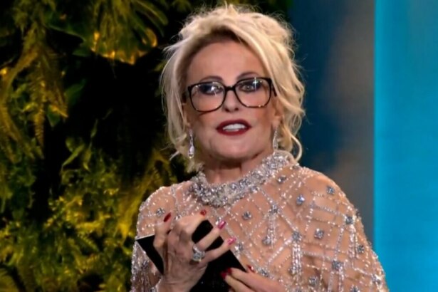 A imagem é um close da apresentadora Ana Maria Braga, com o cabelo loiro preso na final do Chef de Alto Nível. Ela usa um vestido de tule brilhante com pedrarias e gola alta, além de óculos de armação preta. Ela segura um pequeno objeto preto com as duas mãos e parece estar falando, com os lábios pintados de vermelho. O fundo da imagem mostra plantas escuras e uma luz azul.
