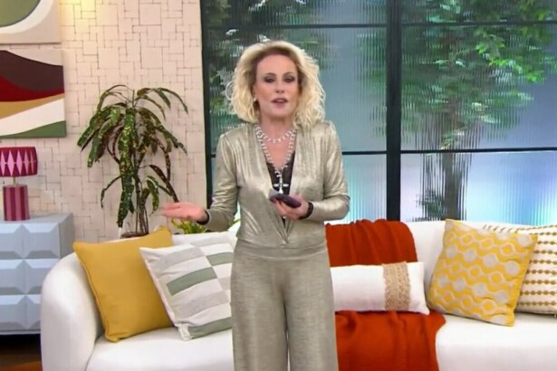 A imagem mostra Ana Maria Braga em pé no cenário do programa "Mais Você". Ela está usando um macacão dourado e brilhante, com um colar prateado de várias voltas. Ela segura um celular em uma das mãos e gesticula com a outra. Ao fundo, um sofá branco com almofadas coloridas, plantas e janelas que simulam um ambiente externo.