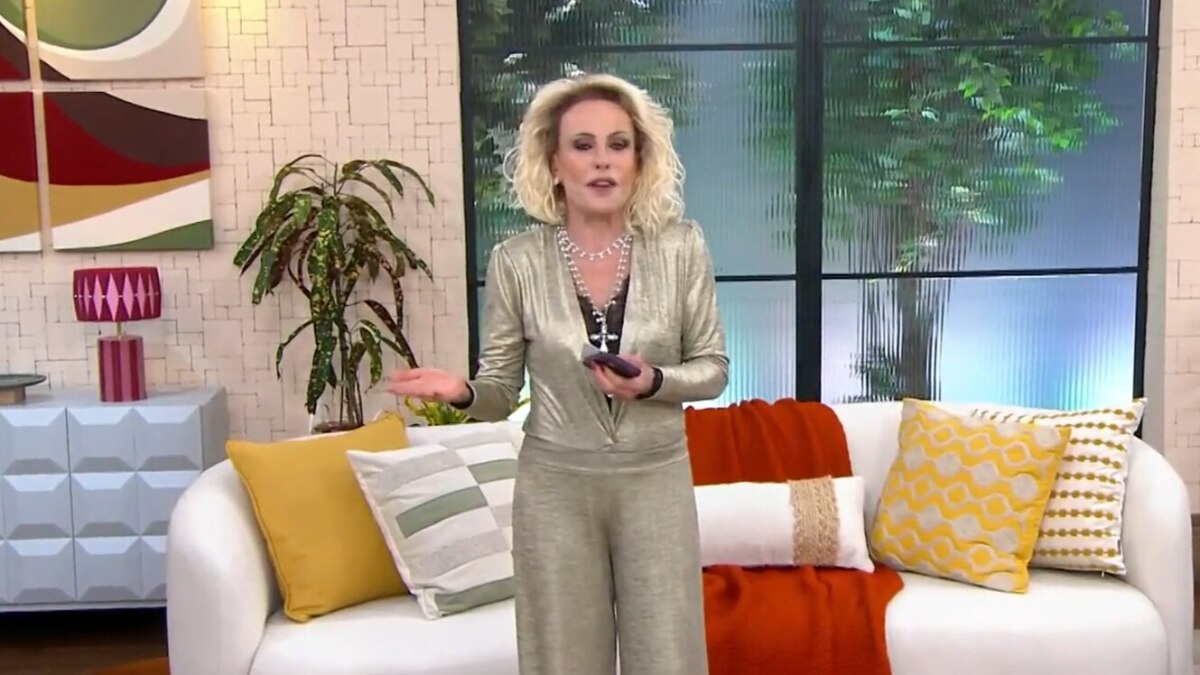 A imagem mostra Ana Maria Braga em pé no cenário do programa "Mais Você". Ela está usando um macacão dourado e brilhante, com um colar prateado de várias voltas. Ela segura um celular em uma das mãos e gesticula com a outra. Ao fundo, um sofá branco com almofadas coloridas, plantas e janelas que simulam um ambiente externo.