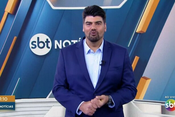 André Azeredo, apresentador do "SBT Notícias", está de pé em um estúdio de televisão, vestindo um terno azul e camisa social clara. Ele tem barba e cabelo escuros e olha para frente, com as mãos cruzadas na altura da cintura. Ao fundo, um telão digital exibe a logomarca "SBT Notícias"