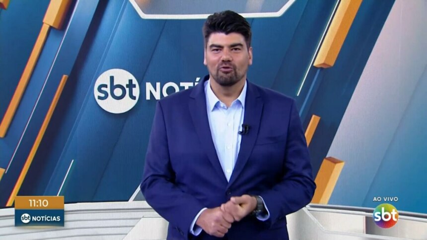 André Azeredo, apresentador do "SBT Notícias", está de pé em um estúdio de televisão, vestindo um terno azul e camisa social clara. Ele tem barba e cabelo escuros e olha para frente, com as mãos cruzadas na altura da cintura. Ao fundo, um telão digital exibe a logomarca "SBT Notícias"