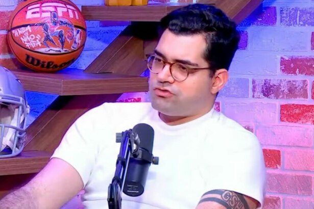 O jornalista Antony Curti em um estúdio de podcast da ESPN. Ele usa óculos e uma camiseta branca, e fala em um microfone com objetos de futebol americano e basquete ao fundo.