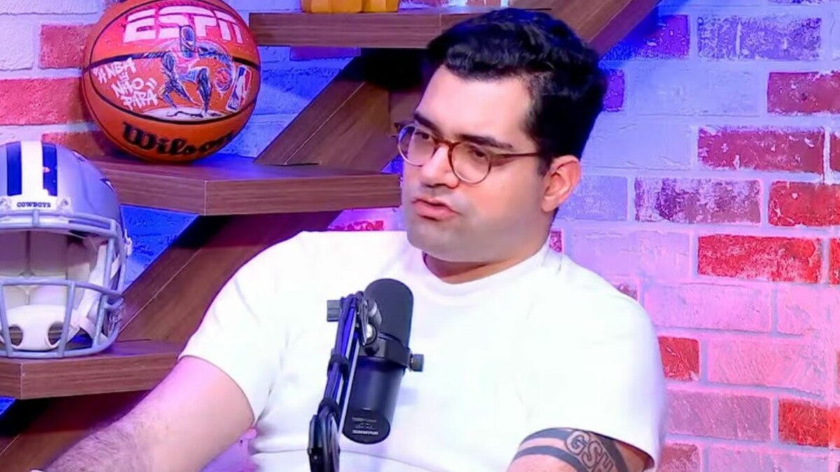 O jornalista Antony Curti em um estúdio de podcast da ESPN. Ele usa óculos e uma camiseta branca, e fala em um microfone com objetos de futebol americano e basquete ao fundo.