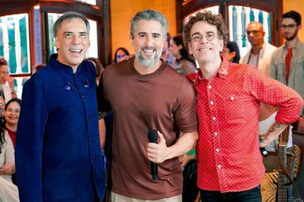 Arnaldo Antunes, Marcos Mion e Nando Reis sorrindo lado a lado, com Mion segurando um microfone, durante o programa Caldeirão.
