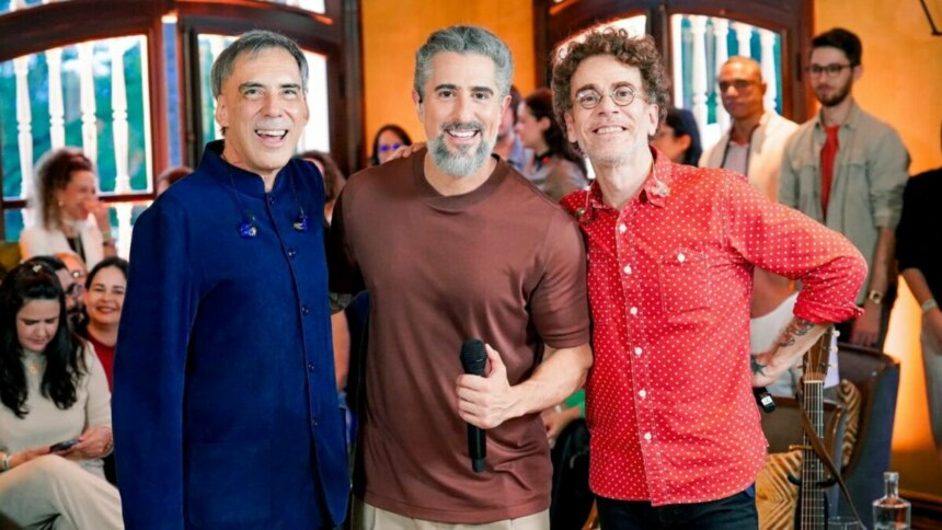 Arnaldo Antunes, Marcos Mion e Nando Reis sorrindo lado a lado, com Mion segurando um microfone, durante o programa Caldeirão.