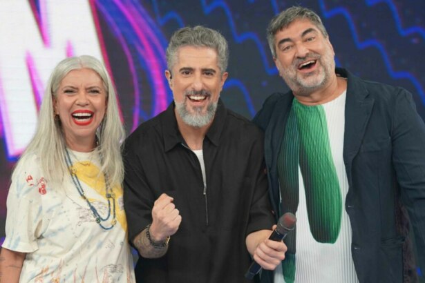 Uma foto de grupo com os apresentadores Astrid Fontenelle, Marcos Mion e Zeca Camargo no cenário do programa 'Caldeirão'. Da esquerda para a direita, estão Astrid de cabelos grisalhos e camiseta estampada, Marcos Mion de camisa preta e Zeca Camargo de jaqueta escura, todos sorrindo para a câmera.