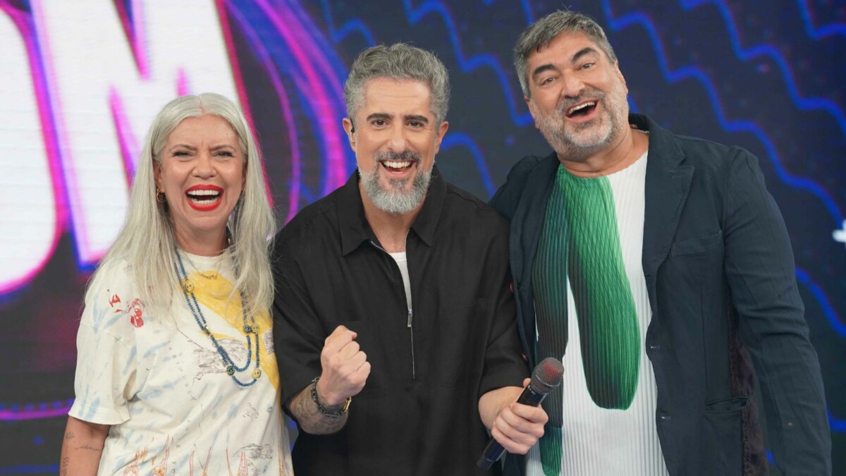 Uma foto de grupo com os apresentadores Astrid Fontenelle, Marcos Mion e Zeca Camargo no cenário do programa 'Caldeirão'. Da esquerda para a direita, estão Astrid de cabelos grisalhos e camiseta estampada, Marcos Mion de camisa preta e Zeca Camargo de jaqueta escura, todos sorrindo para a câmera.