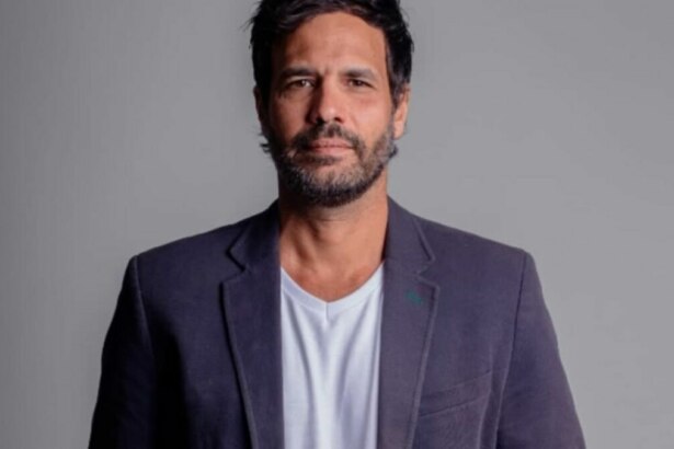 Foto de estúdio do ator Maurício Silveira, com cabelo e barba escuros, vestindo um blazer escuro sobre uma camiseta branca, olhando diretamente para a câmera com uma expressão séria. O fundo é liso e em tom de cinza claro.