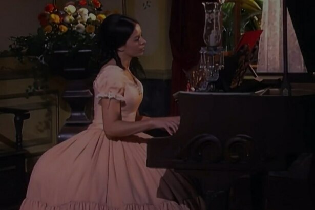 A imagem mostra a atriz Bianca Rinaldi interpretando a personagem "Escrava Isaura". Ela está sentada e toca um piano de madeira escuro. Bianca veste um vestido de época rosa claro, com mangas bufantes. Ao fundo, um vaso de flores, uma cadeira e uma janela escura.