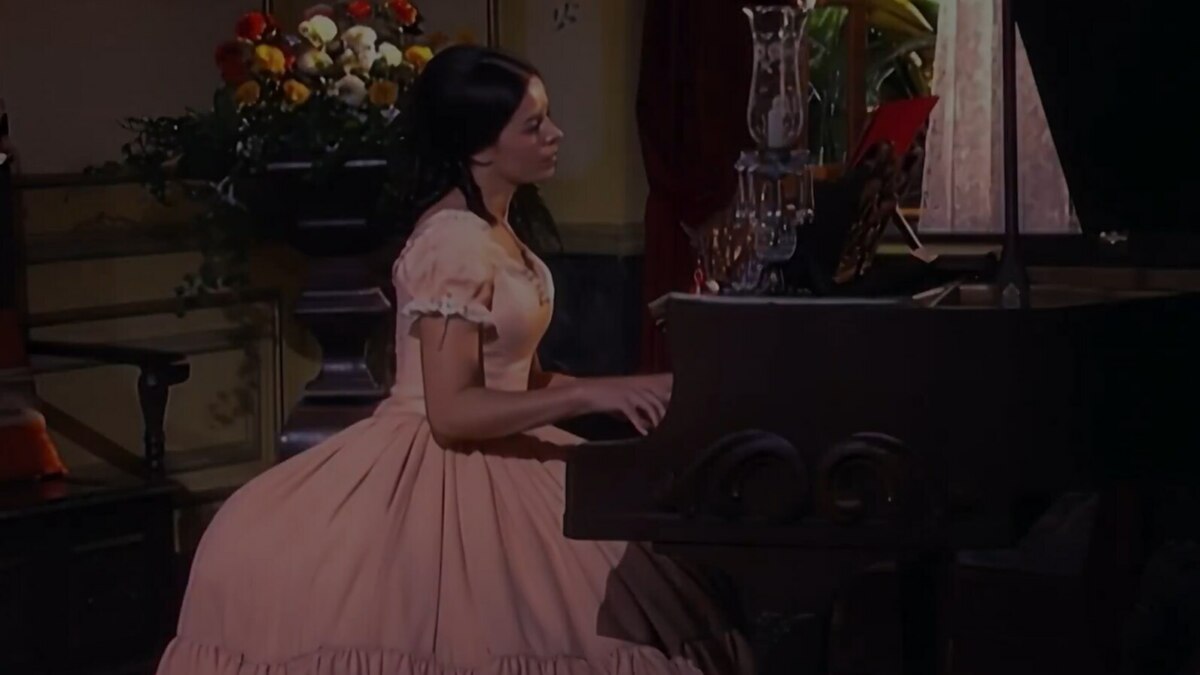 A imagem mostra a atriz Bianca Rinaldi interpretando a personagem "Escrava Isaura". Ela está sentada e toca um piano de madeira escuro. Bianca veste um vestido de época rosa claro, com mangas bufantes. Ao fundo, um vaso de flores, uma cadeira e uma janela escura.
