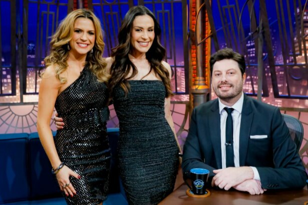 Os apresentadores Danilo Gentili, Beca Milano e Nadja Haddad posam sorrindo juntos no estúdio do programa "The Noite". Danilo está sentado à mesa, e as duas mulheres estão em pé ao seu lado. Beca Milano usa um vestido preto brilhante, e Nadja Haddad usa um vestido preto sem alças.
