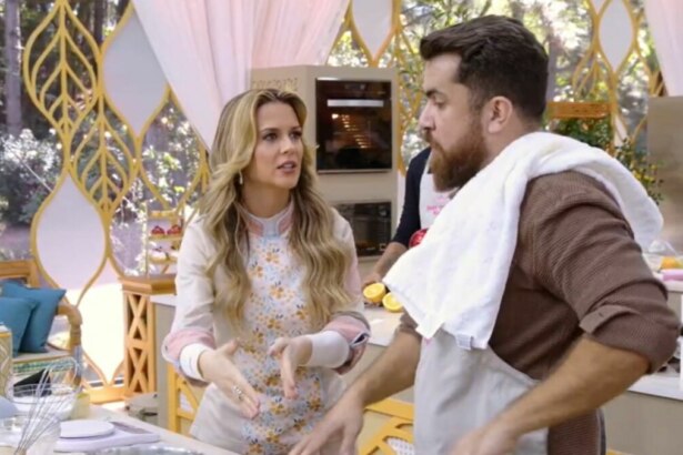 A jurada Beca Milano, do Bake Off Brasil, usando um avental florido, gesticula enquanto conversa com um participante na cozinha do reality.