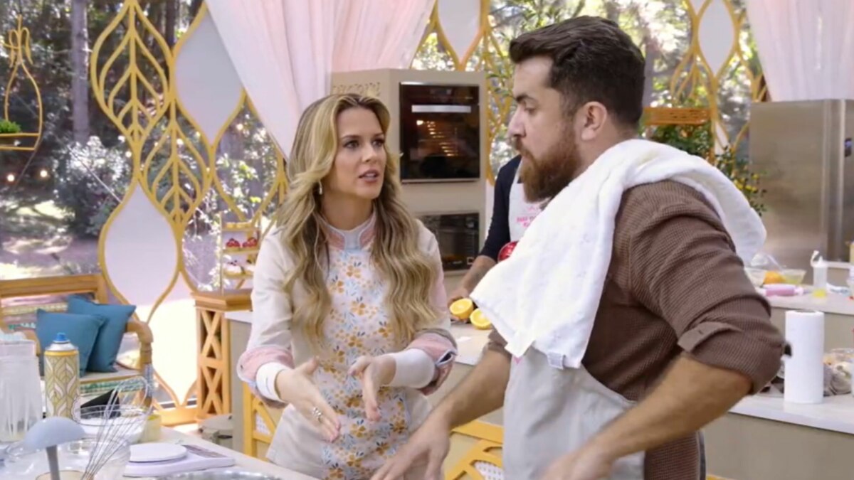 A jurada Beca Milano, do Bake Off Brasil, usando um avental florido, gesticula enquanto conversa com um participante na cozinha do reality.