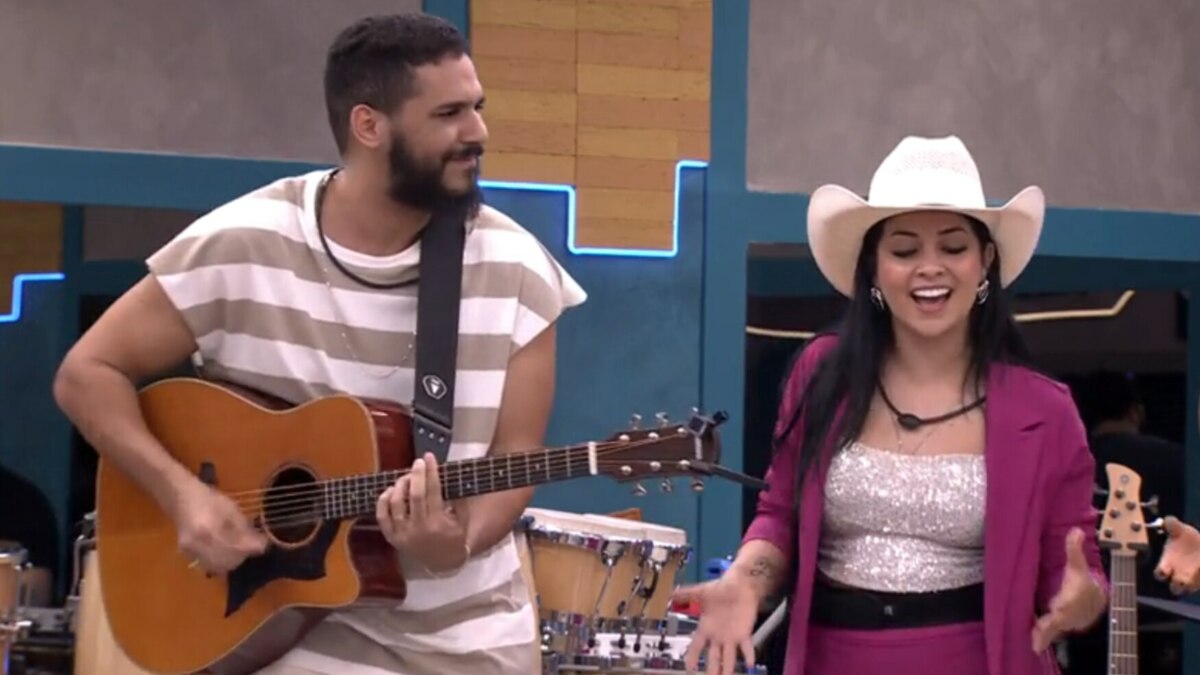 Brenno Casagrande e Biahh Cavalcante no reality Estrela da Casa. Ele toca violão e sorri para ela, que canta usando um chapéu de cowboy e um blazer rosa.