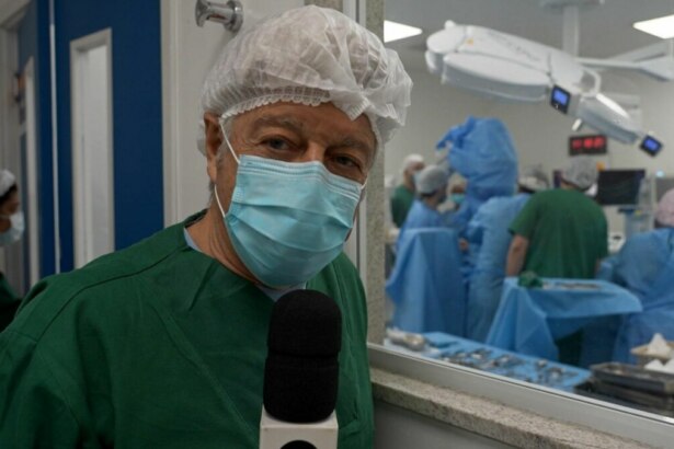 O jornalista Caco Barcellos, em trajes cirúrgicos, máscara e touca, segura um microfone em frente à câmera. Ao fundo, por uma janela de vidro, uma equipe médica trabalha em uma sala de cirurgia, em trecho do Profissão Repórter