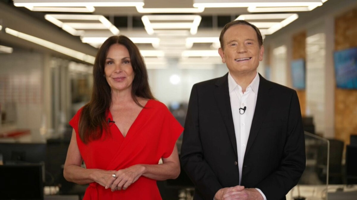 A apresentadora Carolina Ferraz, de vestido vermelho, e o jornalista Roberto Cabrini, de terno preto, estão em um estúdio. Ambos sorriem para a câmera, e o nome do arquivo os associa ao programa 'Domingo Espetacular'.