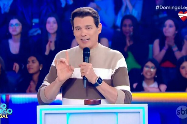 O apresentador Celso Portiolli sorri enquanto fala em um microfone no palco do programa 'Domingo Legal'. Ele usa um suéter listrado de marrom e branco e tem um relógio inteligente no pulso.