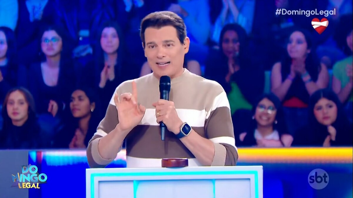 O apresentador Celso Portiolli sorri enquanto fala em um microfone no palco do programa 'Domingo Legal'. Ele usa um suéter listrado de marrom e branco e tem um relógio inteligente no pulso.