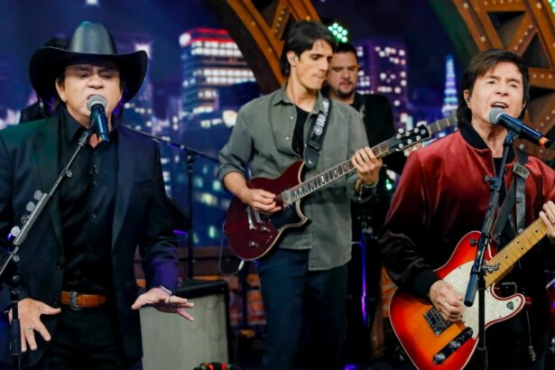 Chitãozinho e Xororó cantando e tocando guitarra no palco do programa The Noite, com a banda ao fundo.