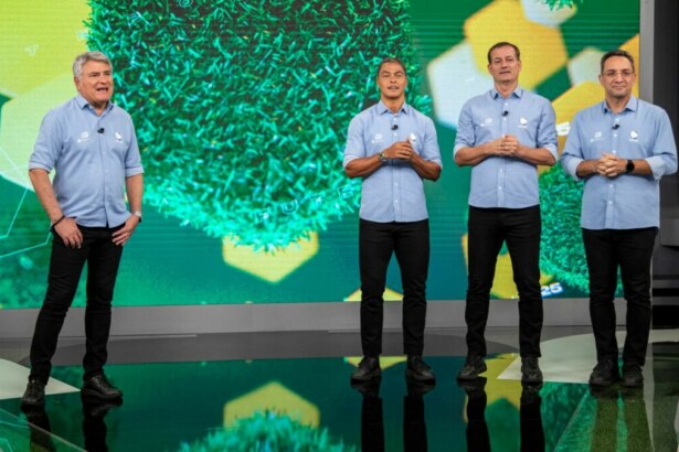 Cleber Machado e sua equipe de comentaristas do Brasileirão na Record posam lado a lado em um estúdio com um fundo temático de gramado e formas geométricas verdes. Da esquerda para a direita: um homem grisalho sorrindo, um homem de camisa polo azul e calça preta com as mãos na frente, Cleber Machado, também de camisa polo azul, e outro comentarista na extrema direita, todos olhando para a frente.