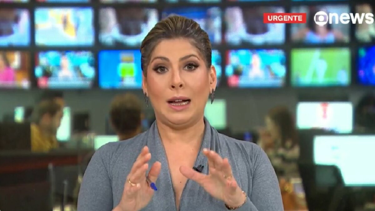 A jornalista Daniela Lima apresentando o jornal da GloboNews, em um estúdio de telejornalismo. Ela fala e gesticula com as mãos. Ao fundo, um painel de telas de TV exibe imagens e o logo da emissora com o aviso "URGENTE". Também é possível ver a redação do canal desfocada.