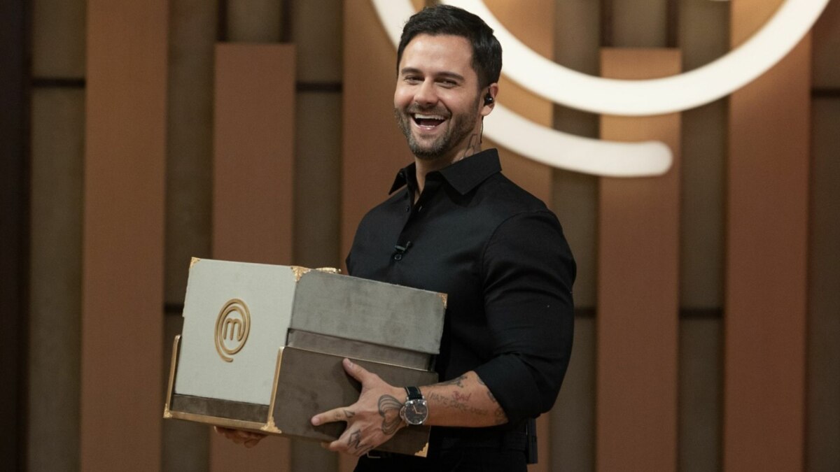 O chef Diego Lozano, de camisa preta, sorri para a câmera enquanto segura uma caixa com o logo do 'MasterChef'. Ele está no cenário do programa.