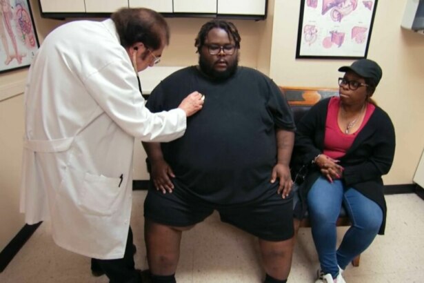 A imagem mostra uma cena do programa "Quilos Mortais". O Dr. Nowzaradan, com seu jaleco branco, está examinando o paciente Travis com um estetoscópio. Travis, que é um homem negro com sobrepeso, está sentado em um banco e veste uma camiseta e shorts pretos. Ao seu lado, a esposa dele o acompanha, com uma expressão séria. Ela está vestindo uma blusa vermelha, casaco preto, calça jeans e um boné preto. O fundo da imagem é um consultório médico, com armários e quadros.