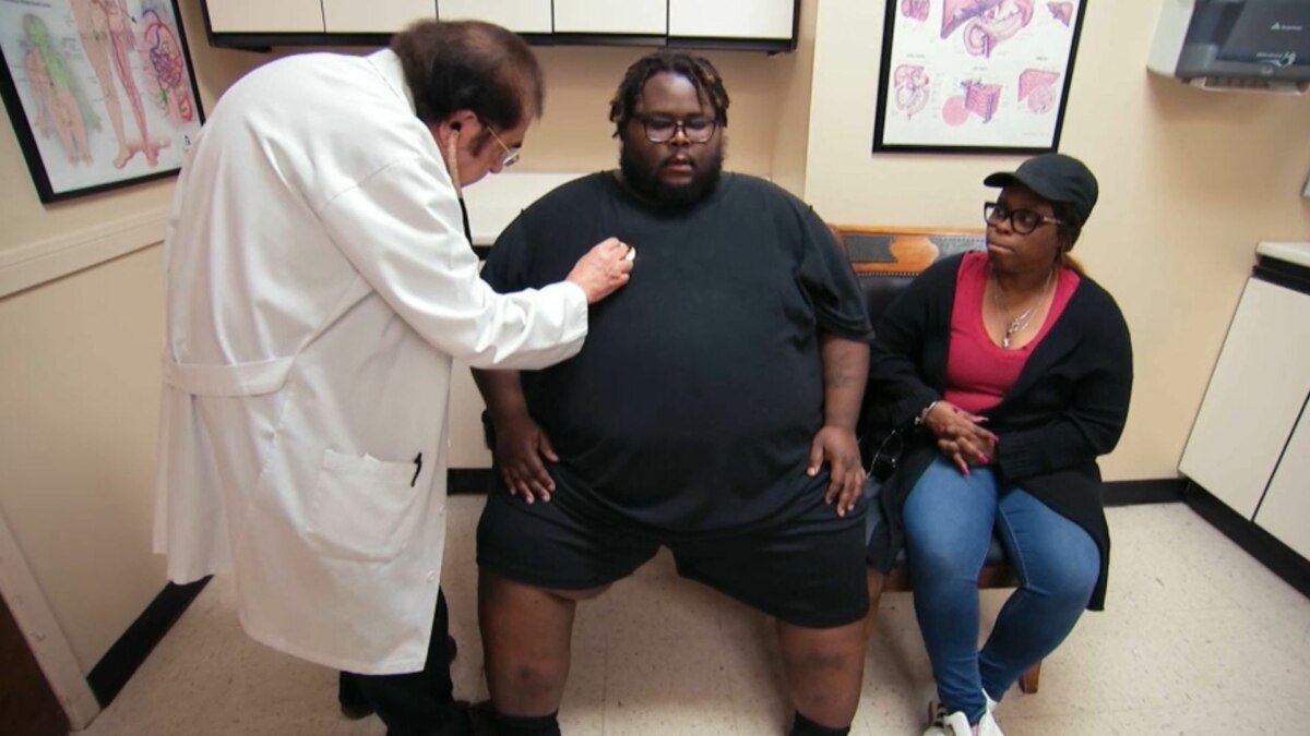 A imagem mostra uma cena do programa "Quilos Mortais". O Dr. Nowzaradan, com seu jaleco branco, está examinando o paciente Travis com um estetoscópio. Travis, que é um homem negro com sobrepeso, está sentado em um banco e veste uma camiseta e shorts pretos. Ao seu lado, a esposa dele o acompanha, com uma expressão séria. Ela está vestindo uma blusa vermelha, casaco preto, calça jeans e um boné preto. O fundo da imagem é um consultório médico, com armários e quadros.