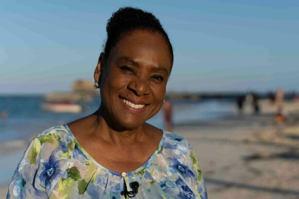 A jornalista Dulcineia Novaes sorri para a câmera em uma praia da República Dominicana, em trecho do Globo Repórter. Ela tem os cabelos presos e usa uma blusa florida de tons azuis e brancos. Ao fundo, um mar azul e areia clara com um barco distante.
