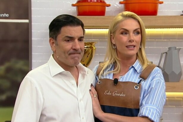 Os apresentadores Edu Guedes e Ana Hickmann em um estúdio de TV, no retorno do quadro "Fica com a Gente". Edu Guedes, de camisa branca, tem uma expressão pensativa. Ana Hickmann, com a mão em seu ombro, o observa. Ela usa um avental com o nome dele bordado.