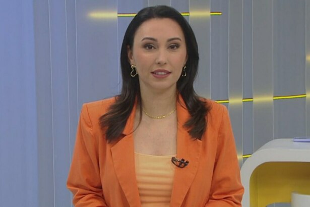 A imagem é um close da apresentadora Eveline Poncio em um estúdio de TV. Ela sorri levemente e olha para a câmera. Eveline usa um blazer laranja sobre uma blusa de cor pêssego, um colar dourado e brincos com formatos de coração. Ao fundo, um cenário claro de televisão e uma caneca com a hashtag "#FazParte" podem ser vistos.