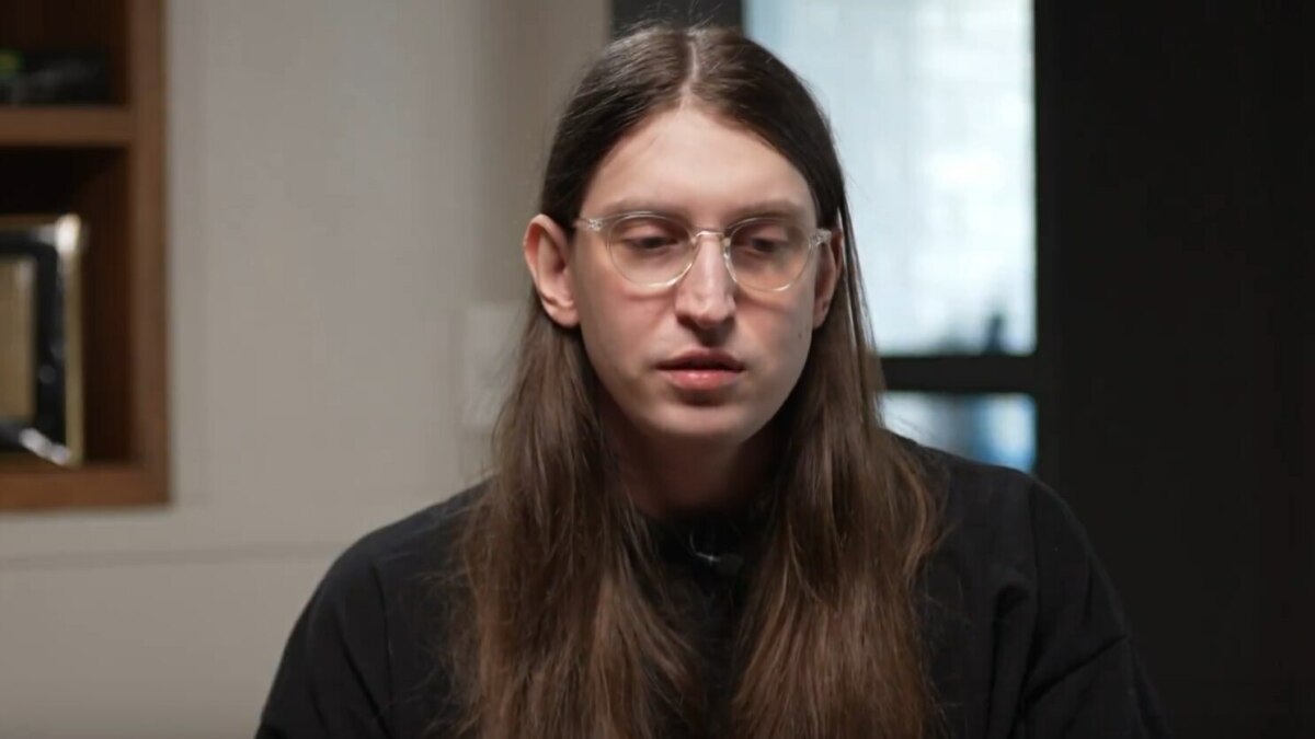 O youtuber Felca, de cabelo comprido e óculos, olha para baixo com uma expressão séria. Ele está em um ambiente interno, em trecho de entrevista ao Fantástico