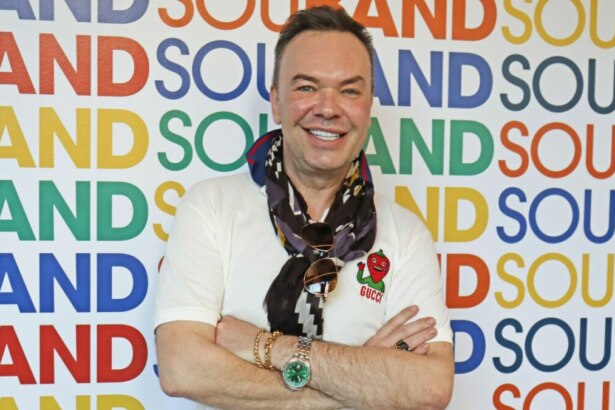 A imagem mostra Felipeh Campos em pé, sorrindo e olhando para a câmera. Ele está de braços cruzados e usa uma camiseta branca com um emblema da Gucci, um lenço no pescoço, pulseiras e um relógio verde. Ao fundo, um painel repete a palavra "BAND" em cores variadas.