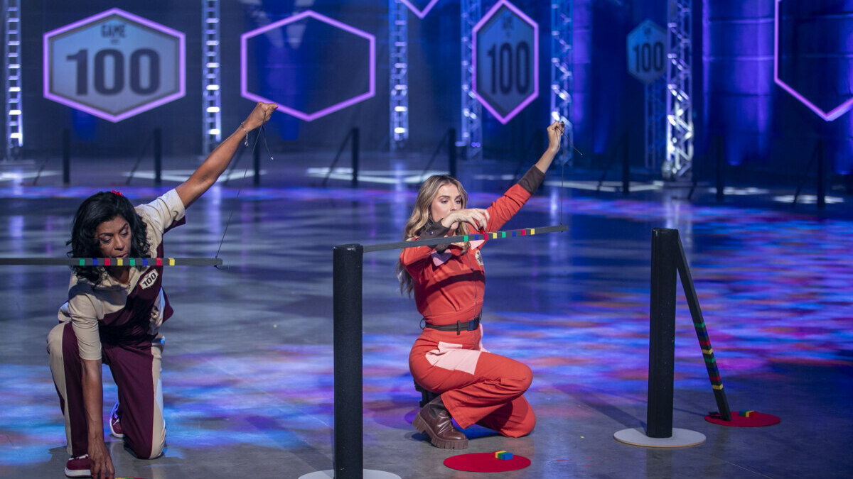 Márcia Fu e Rafa Brites equilibram peças de dominó em prova do Game dos 100, ajoelhadas no cenário iluminado