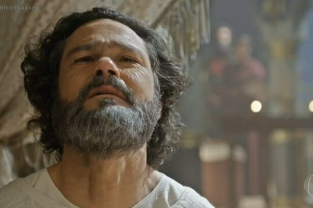 A imagem é um close do ator Gabriel Gracindo em uma cena da novela "O Rico e Lázaro", onde ele interpreta o profeta Daniel. Ele tem o cabelo e a barba grisalha e está olhando para cima com uma expressão séria. Ao fundo, o cenário da novela e uma figura desfocada podem ser vistos.