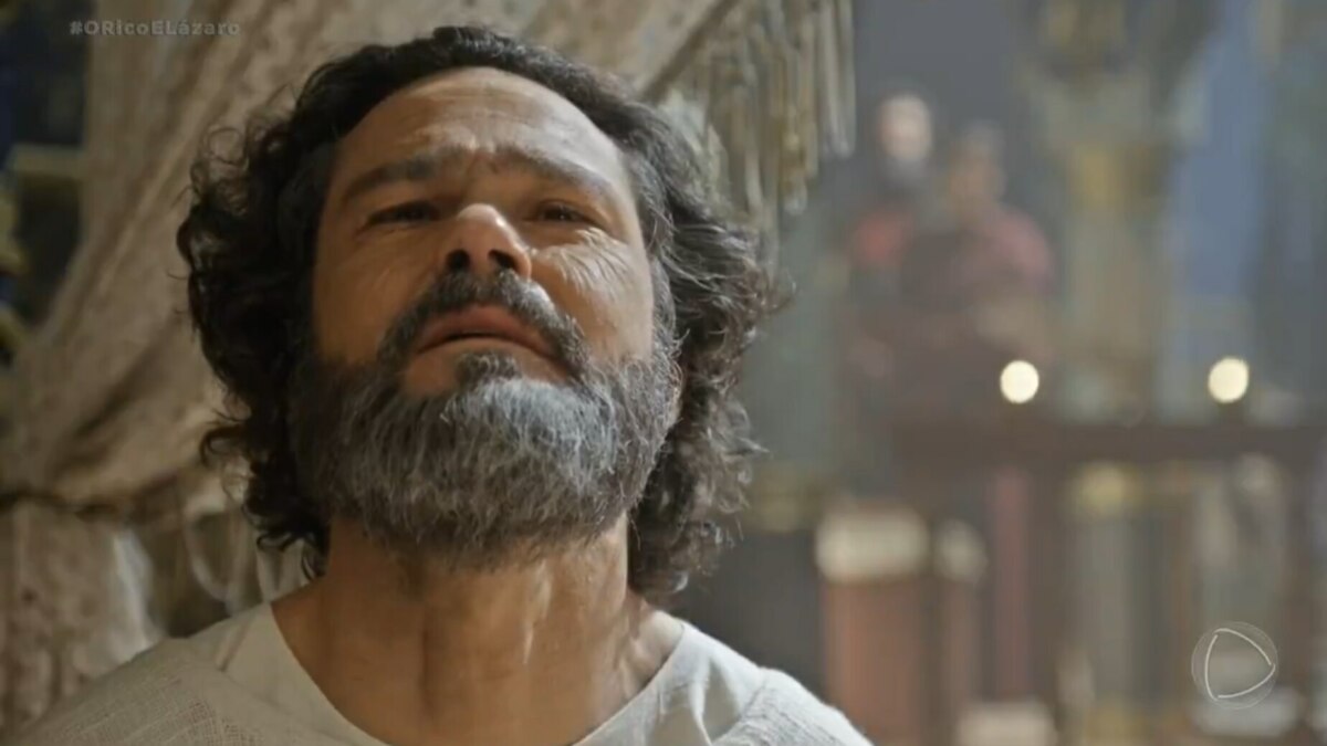 A imagem é um close do ator Gabriel Gracindo em uma cena da novela "O Rico e Lázaro", onde ele interpreta o profeta Daniel. Ele tem o cabelo e a barba grisalha e está olhando para cima com uma expressão séria. Ao fundo, o cenário da novela e uma figura desfocada podem ser vistos.