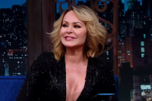 A atriz Gabriela Spanic, com cabelo loiro na altura dos ombros, sentada em um sofá azul em um estúdio de televisão. Cotada para A Fazenda 17, ela está sorrindo e olhando para o lado, vestindo um casaco preto brilhante e decotado. O fundo é um painel digital que simula uma vista noturna de uma cidade, com luzes de prédios.