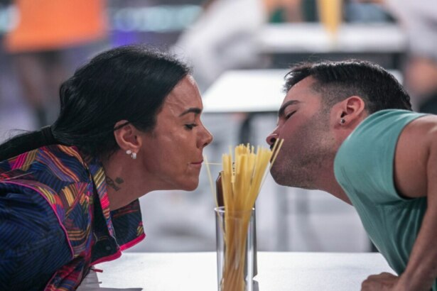 Uma mulher de cabelos escuros presos e um homem com barba e camiseta azul clara se inclinam um em direção ao outro, tentando comer um espaguete comprido que está em um copo alto no centro, durante a "Prova do Macarrão na Lata" no terceiro episódio do "Game dos 100". Ambos estão com a boca perto do macarrão, concentrados na tarefa, em um ambiente de estúdio