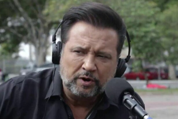 A imagem é um close do jornalista Geraldo Luís durante uma reportagem para o programa "Aqui Agora". Ele está com uma expressão séria, falando em um microfone. Geraldo usa um fone de ouvido preto e uma camisa preta. O fundo está desfocado, mostrando uma rua com árvores e pessoas.