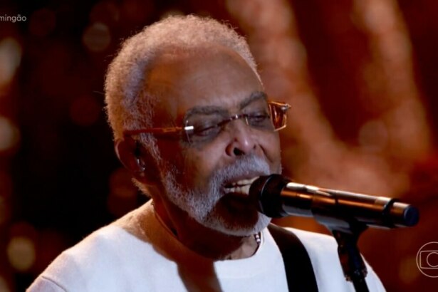 O cantor Gilberto Gil, usando óculos e um suéter branco, canta com os olhos fechados em um microfone durante uma apresentação no palco do Domingão.
